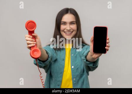 Portrait of delighted smiling woman holding out retro phone and cell phone, choose best device for you, wearing casual style jacket. Indoor studio shot isolated on gray background. Stock Photo