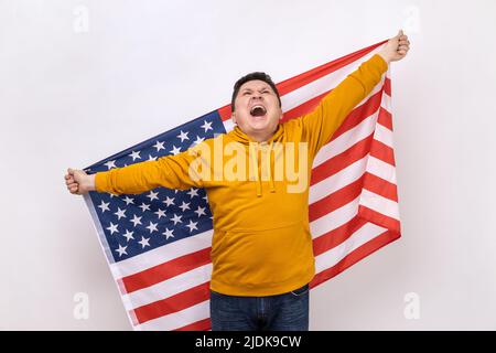 Portrait of middle aged man holding USA flag and screaming happily, looking up, celebrating national holiday, wearing urban style hoodie. Indoor studio shot isolated on white background. Stock Photo