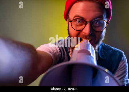 Smiling hipster man with beard screaming at megaphone, protesting while livestreaming, wants to be heard POV, wearing beanie hat. Indoor studio shot isolated on colorful neon light background. Stock Photo