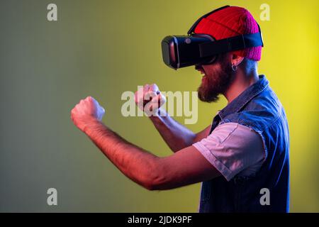 Side view of hipster man in virtual reality glasses on head playing fighting game, holding clenched fists up ready to boxing. Indoor studio shot isolated on colorful neon light background. Stock Photo
