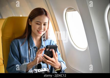 Beautiful millennial asian female passenger sits at the window seat using her smartphone during the flight. Stock Photo