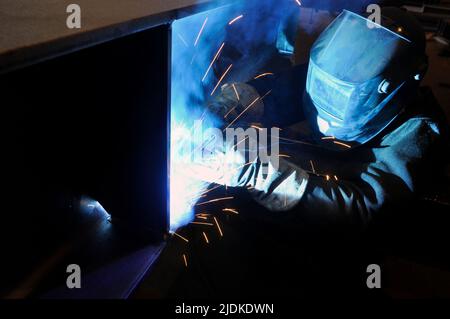 A welder in protective overalls and a welding mask welds metal structures by welding in a hard-to-reach place. forced posture. Stock Photo