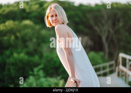 Side view of middle-aged woman leaning on railing of bridge in forest, luxuriating in sunshine, looking at camera.  Stock Photo