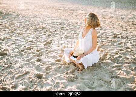Middle-aged woman sitting with crossed legs on sandy beach on sunny day, putting hands on straw hat, looking at sun.  Stock Photo