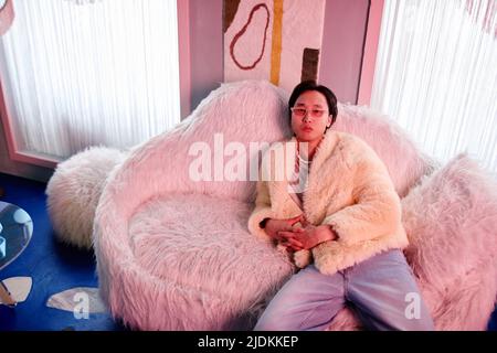 Portrait of Asian young man dressed in fur coat and party outfit relaxing in clu lounge lit by pink neon lights Stock Photo