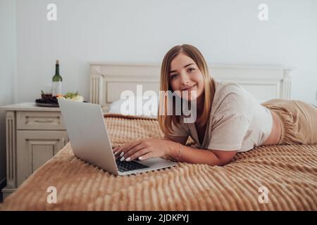 Cheerful Caucasian Woman Laying on the Bed and Looking Into Camera While Working on Laptop, Freelancer at Work Stock Photo
