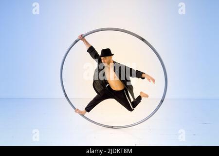 Full body of active male acrobat performing trick with cyr wheel while standing on one leg with raised arm against light background Stock Photo