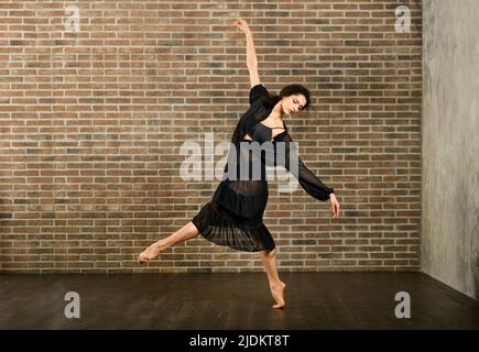 Full body of barefoot flexible female ballet dancer standing on one leg while rehearsing dance against brick wall in studio Stock Photo