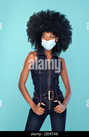 Happy African American female in trendy denim clothes and face mask looking away while standing against turquoise background during pandemic Stock Photo