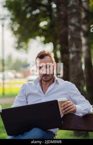 Concentrated man works in the park with a laptop, drinks coffee. A young man on a background of green trees, a hot sunny summer day. Warm soft light, close-up. Stock Photo