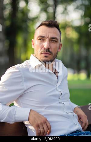 A serious business man in a white shirt is resting while sitting on a bench. Young man in city park, warm sunny summer day. Warm soft light, close-up. Stock Photo