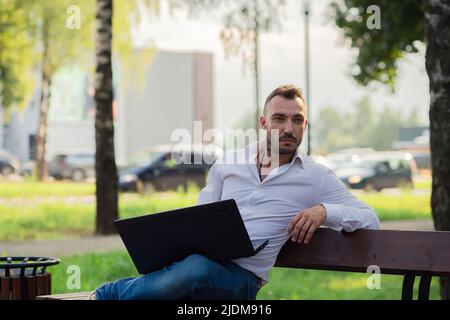 Thoughtful business man in a white shirt is working in the park with a laptop. Young man on the background of passing cars, hot sunny summer day. Warm soft light, close-up. Stock Photo