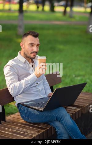 A man looking at the camera is working in the park with a laptop, drinking coffee. A young man on a background of green trees, a hot sunny summer day. Warm soft light, close-up. Stock Photo