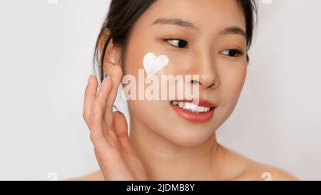 Facial skincare concept. Young korean lady with heart-shaped face cream on cheek posing isolated on white background Stock Photo