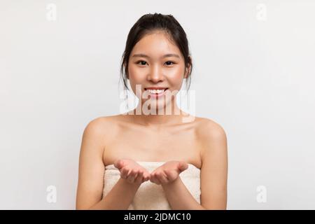 Happy young asian lady wrapped in bath towel demonstrating something on empty palms, showing invisible object Stock Photo