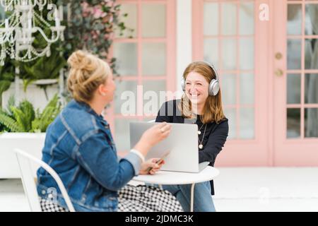 Two smiling women sitting at table with laptop and phone wearing headphones Stock Photo