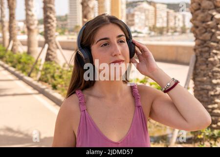 Young woman in a pink dress, holds and listens to music on her black headphones on a sunny day in the city. Stock Photo