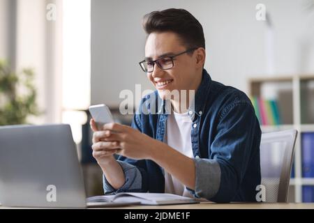 Smiling Young Male Entrepreneur Using Smartphone While Sitting At Desk With Laptop Stock Photo