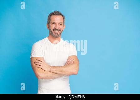 Middle aged grey haired man with standing with arms folded happy smiling on camera wearing white t-shirt isolated on blue background. Mature fit man, healthy lifestyle concept. Stock Photo