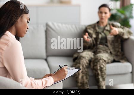 Black Female Psychotherapist Taking Notes During Therapy Session With Military Woman Stock Photo