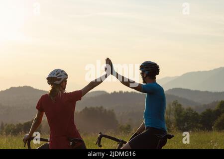 Two road cyclists silhouettes celebrating a ride finish with a high five greeting, on amazing mountain view at sunset Stock Photo