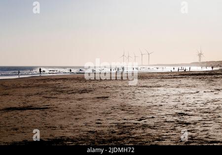 The beach at sunset with people and wind turbine power generators silhouetted by the backlight of sunlight. Necochea, Argentina. Stock Photo