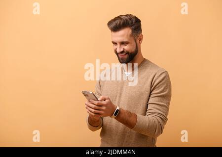 Smiling young bearded man using mobile phone isolated by beige background, 30s guy texting online, scrolling news feed, web surfing on the mobile phone, enjoying chatting in social medias Stock Photo