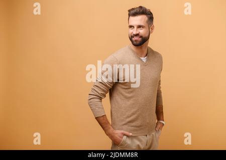 Portrait of happy satisfied bearded young man in pullover sweater standing and looking away with light smile isolated on beige. Indoor studio shot of handsome guy Stock Photo