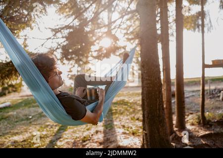 Attractive young man resting in hammock with coffee, tea in hammock in forest. Rear. Stock Photo