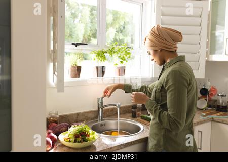 Biracial young woman in hijab washing vegetables at sink in kitchen Stock Photo