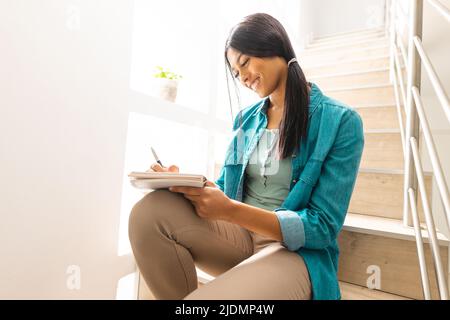 Smiling biracial young woman writing in book while sitting on stairs at home Stock Photo