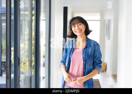 Portrait of smiling asian young woman with short hair wearing denim jacket standing by window Stock Photo