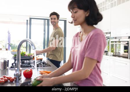 Smiling caucasian young man looking at asian girlfriend cleaning vegetables in sink in kitchen Stock Photo