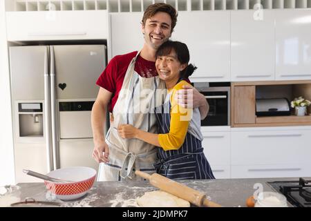 Portrait of cheerful multiracial young couple with messy faces embracing while kneading dough Stock Photo
