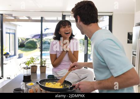 Caucasian young man talking with cheerful asian girlfriend while making scrambled eggs in kitchen Stock Photo