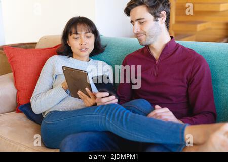 Multiracial young couple using mobile phone and digital tablet while sitting on sofa in living room Stock Photo