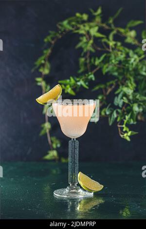 Glass of refreshing Pegu Club cocktail served on dark green table surface surround of fresh lime, shallow depth of the field. Stock Photo