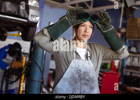 Confident mid adult asian female welder wearing protective worker while working in workshop Stock Photo