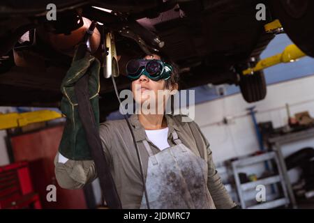 Mid adult asian female welder using work tools while wielding car under car lift in workshop Stock Photo
