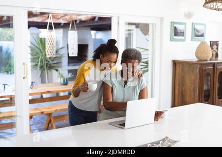 African american young woman with coffee cup standing by mother using laptop on table at home Stock Photo