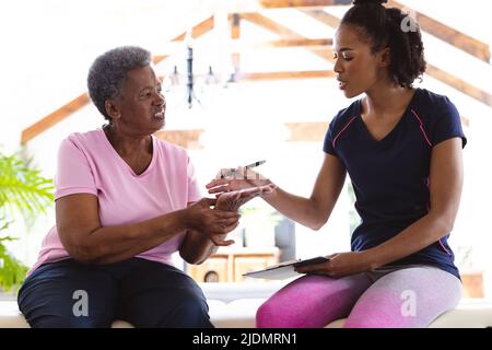 African american senior woman showing hand to female physiotherapist while sitting on table at home Stock Photo