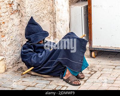 A side view of homeless beggar man sitting outdoors, leaning against a ...