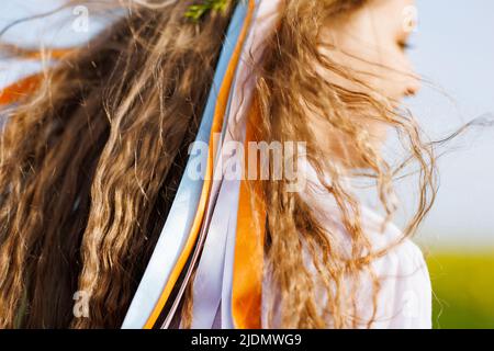 Light brown wavy hair of cheerful teenage girl decorated with bright Ukrainian summer wreath with yellow blue wild flowers and long multi-colored ribb Stock Photo