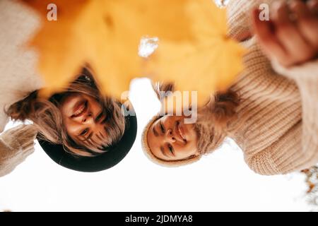 Two charming young women dressed in knitted sweaters are photographed from below against background of autumn sky. Leaf fall. Positive emotions Stock Photo