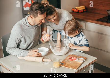 Young couple, man and woman Teaches son 4 - 5 years to Bake cookies, knead dough, work with flour in kitchen at home, happy hours of young family Stock Photo