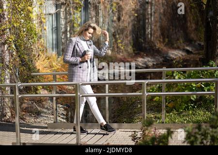 Attractive young woman with long blonde hair in stylish clothes walks along wooden bridge in city park with cup of coffee in her hands . Autumn mood Stock Photo