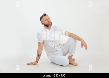 Bearded barefoot thoughtful man with tattoo sitting on the floor in white outfit on white background. Business thoughts Stock Photo