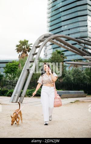 Young woman talking on a phone and walking with her dog on a urban park Stock Photo