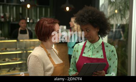 Female manager explaining small business Coffee shop to new employee. Two diverse employees wearing aprons standing inside cafe Stock Photo
