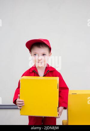 boy 3-4 years old in a red uniform and cap with a yellow cardboard box in his hands. express delivery of happiness. positive feedback. place for adver Stock Photo